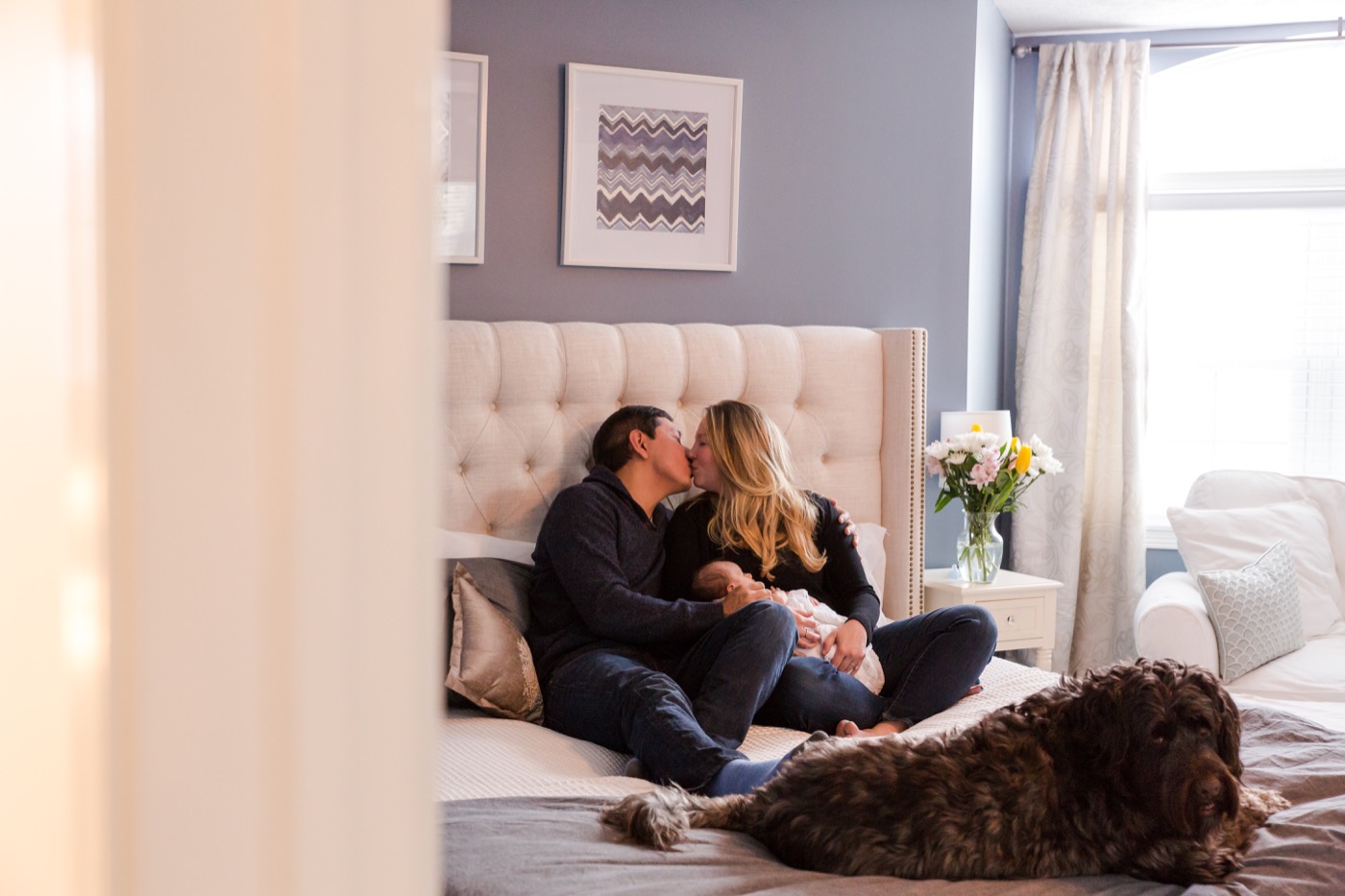 New parents sitting together on a bed holding their newborn baby during an in-home newborn photography session in Grand Rapids Michigan — Jules K Photography