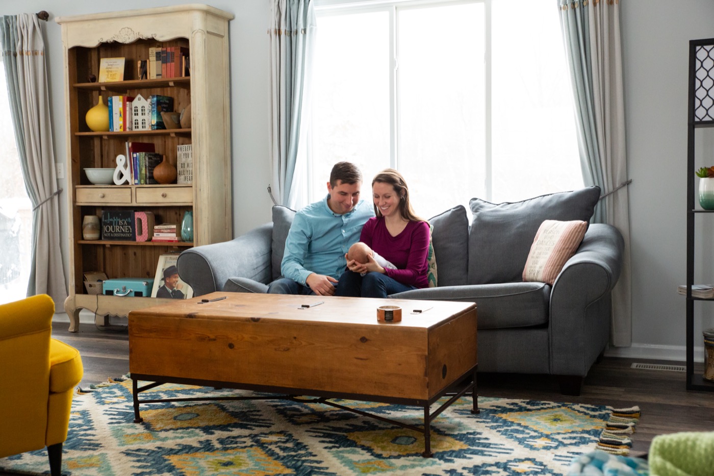 Parents sitting together on a couch in their living room holding their newborn baby during an in-home newborn photography session in Grand Rapids Michigan — Jules K Photography
