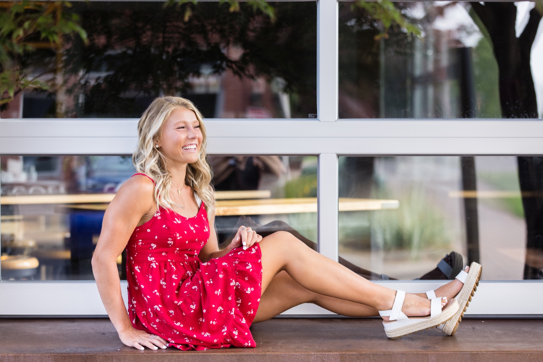 Senior girl in red dress sitting against glass storefront in downtown Holland Michigan
